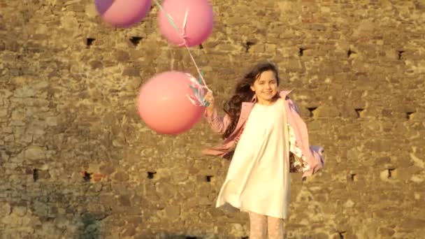 kız beyaz bir elbise ve pembe bir ceket güneşin Ortaçağ duvarlar zemin karşı parlak çok renkli balonlar ile - Video, Çekim