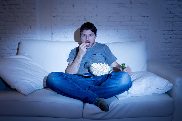 young television addict man sitting on home sofa watching TV eating popcorn and drinking beer bottle - Fotó, kép