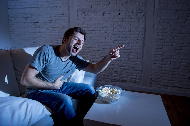 young television addict man sitting on home sofa watching TV and eating popcorn laughing crazy - Fotó, kép