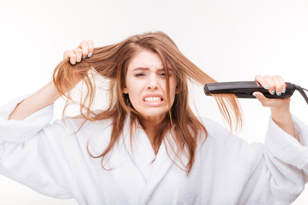 Angry irritated young woman straightening her hair using straightener  - Foto, afbeelding