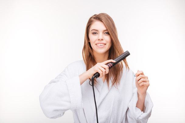 Beautiful happy young woman in bathrobe using hair straightener - Foto, Imagem