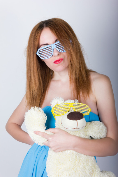 girl in a blue dress and sunglasses in the style of disco, hugging a teddy bear in the same glasses - Fotoğraf, Görsel