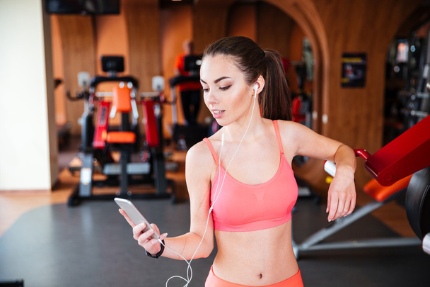 Smiling woman athlete with earphones using smartphone in gym - Photo, Image