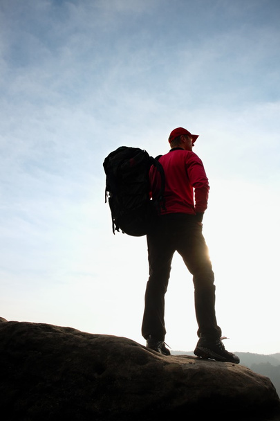 Один турист с большим рюкзаком стоять на краю скалы и смотреть в глубокую долину ниже
.  - Фото, изображение