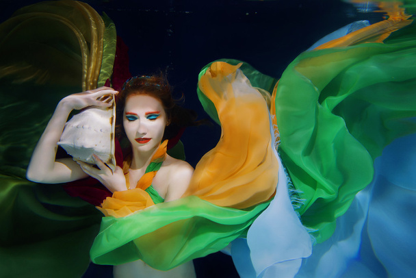 水中の暗い背景にカラフルな服に大きな貝殻と赤い髪の少女 - 写真・画像