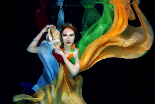 Red hair girl with big sea shell in colorful clothes on the dark background underwater - Photo, Image