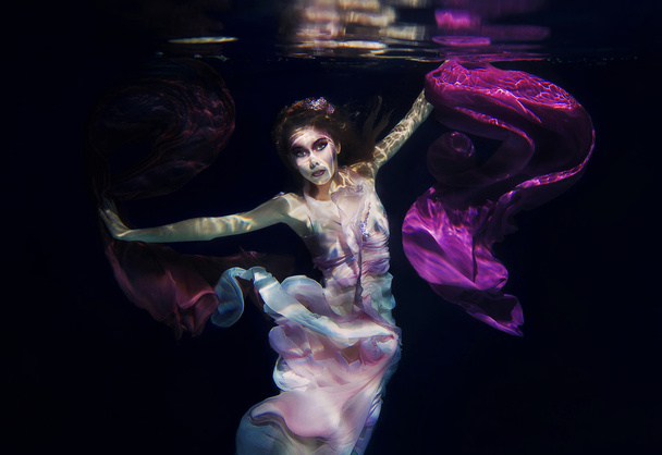 Mädchen in bunten Kleidern auf dem dunklen Hintergrund schwimmt unter Wasser - Foto, Bild