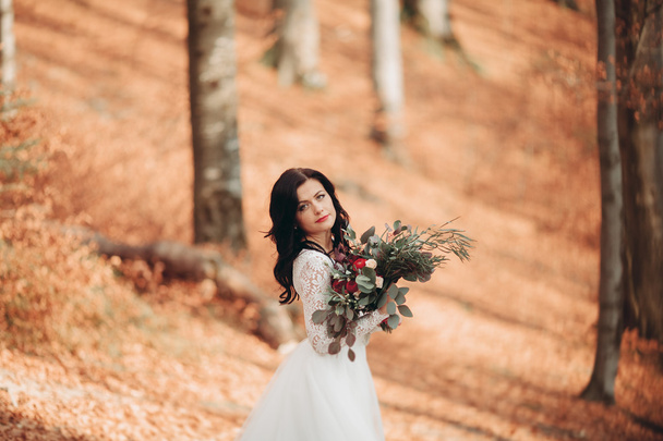 Πανέμορφη μελαχρινή νύφη σε κομψό φόρεμα, κρατώντας την ανθοδέσμη που παρουσιάζουν κοντά στο δάσος και τη λίμνη - Φωτογραφία, εικόνα