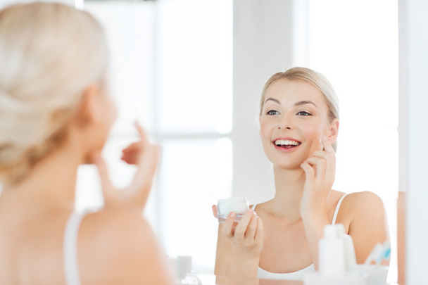 happy woman applying cream to face at bathroom - Foto, Imagen