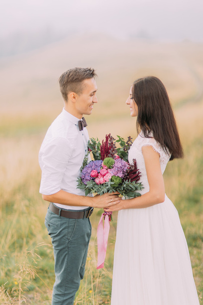 Happy bride and groom with bouquet in hands lovingly look at each other. Misty field on background - Фото, зображення