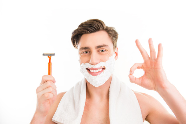 Attractive happy  man with towel, foam on his face and razor ges - Fotó, kép