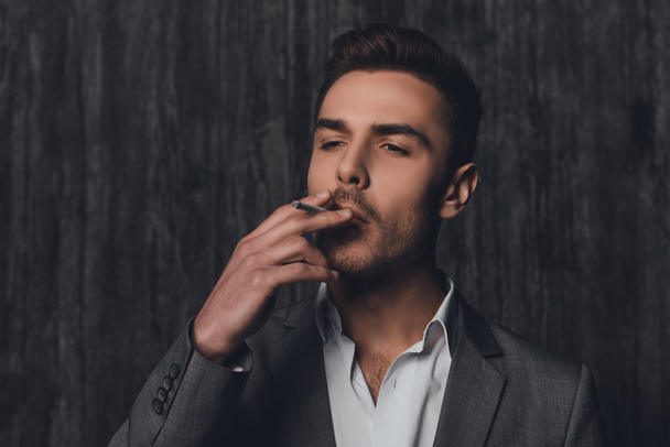 Handome brutal man in suit on the gray background smoking a ciga - 写真・画像