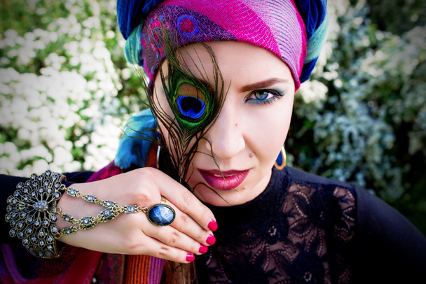 portrait of the beautiful girl in a turban with a peacock feather in her hands on the flowers of spirea background - Foto, afbeelding