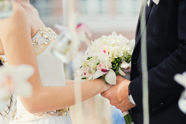 Las manos de la novia y el novio se cierran en la ceremonia de boda cerca del arco de la boda. Flores de orquídea ramo de novia en sus manos
 - Foto, Imagen