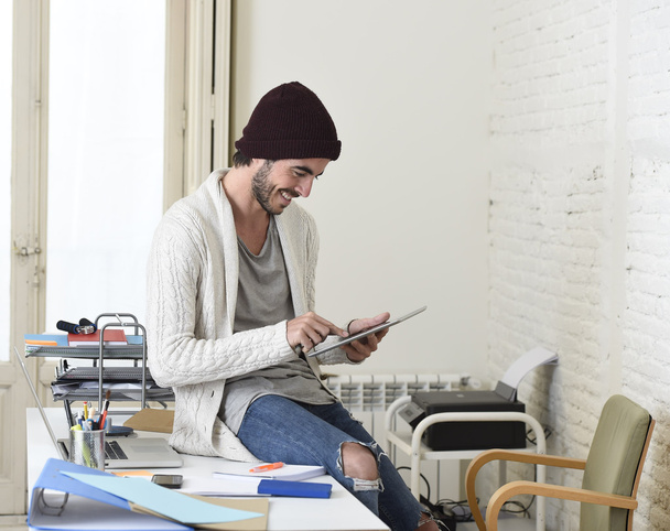 young trendy businessman in beanie and cool hipster informal look sitting on home office desk using digital tablet pad happy - 写真・画像