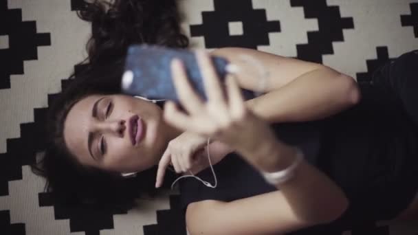 Mladá a krásná dívka leží na podlaze, poslouchá hudbu prostřednictvím smartphone a dělá lipsync. - Záběry, video
