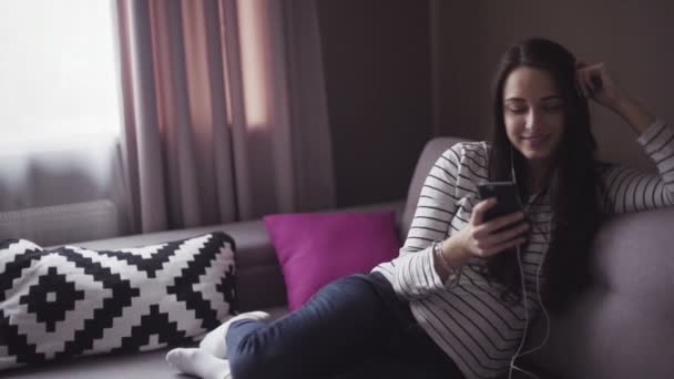 Chica morena joven está sentado en una sala de estar en un sofá y escuchar la música en el dispositivo móvil
. - Metraje, vídeo