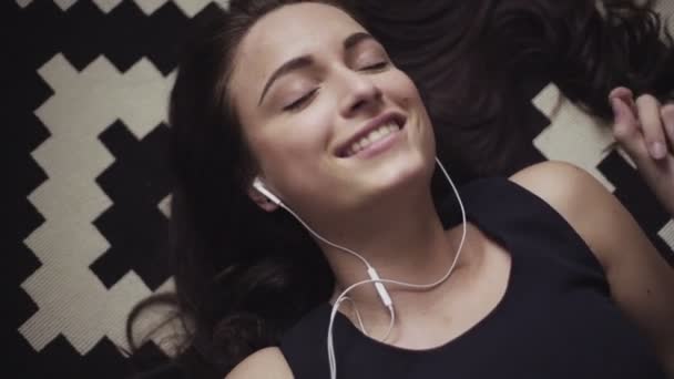 Панорамне зображення того, як молода жінка лежить на підлозі, слухає і переходить до музики на своєму мобільному пристрої з підключеними навушниками
. - Кадри, відео