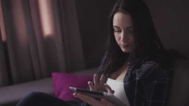 Handsome brunette girl sits on a couch indoors and uses tablet for business. - Video, Çekim