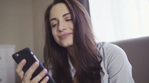 Slow motion beelden hoe brunette meisje zit op een bank in een woonkamer en kijken naar smartphone scherm. - Video
