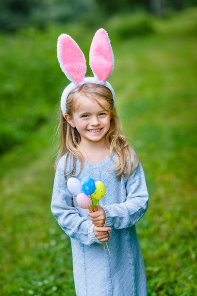 Portrait of smiling little girl with long blond hair wearing rabbit or bunny ears and blue knitted dress and holding bunch of painted colour eggs on sunny day in spring park. Easter celebrations - Φωτογραφία, εικόνα