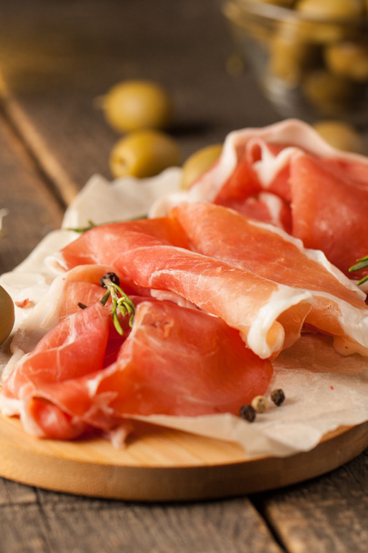 closeup of thin slices of prosciutto with mixed olives and paprika on wooden cutting board - Photo, Image