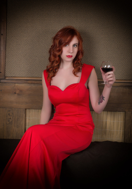 Elegant young redhead woman in a red dress, having a glass of red wine - Zdjęcie, obraz