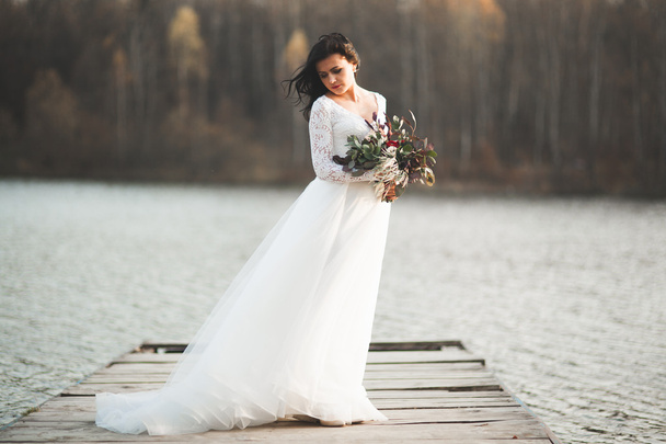 Gorgeous brunette bride in elegant dress holding bouquet  posing near forest and lake - 写真・画像