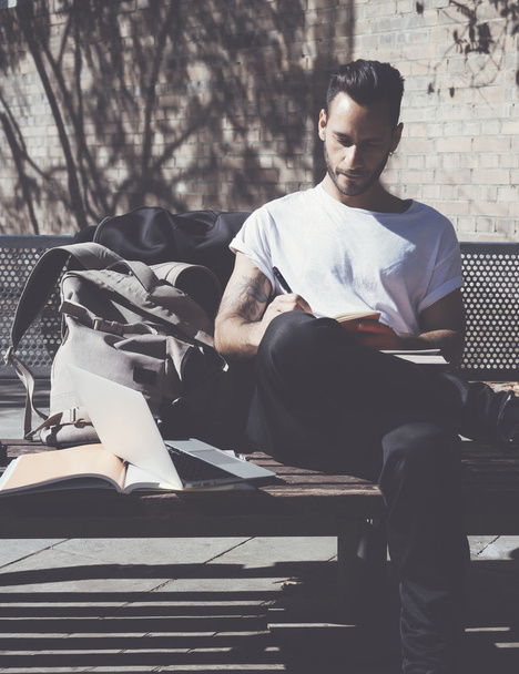 Bearded student wearing white tshirt sitting city park and draws notepad. Studying at the University, working project. Books, generic design laptop, backpack bench.  Vertical - Foto, Imagem
