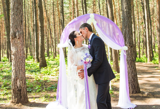 maravilloso elegante novia feliz y novio de pie en una ceremonia de boda en el jardín verde cerca del arco púrpura con flores
 - Foto, Imagen