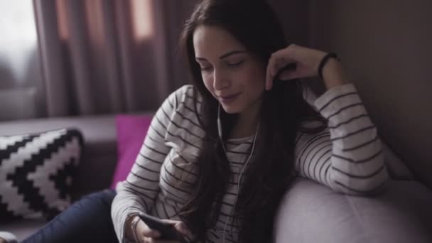 Young woman is sitting on a couch in a living room, girl listens to the music from her mobile device through earphones. - Video, Çekim