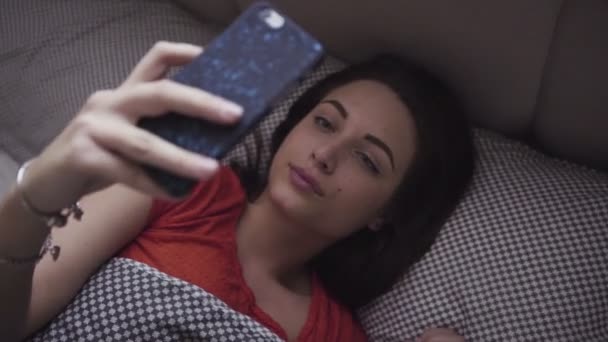 Mladá žena leží na posteli a její mobilní zařízení. Dívka při pohledu na obrazovku smartphone a reaguje na něco, s úsměvem. - Záběry, video