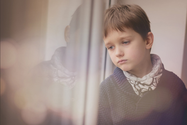 Szomorú átgondolt kis fiú néz az ablakon keresztül. - Fotó, kép