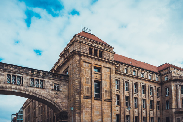 Низкоповоротный архитектурный экстерьер старого здания с коричневым каменным фасадом в классическом стиле с переходом через арочный пешеходный мост, обрамленный синим небом с тяжелыми белыми облаками
 - Фото, изображение