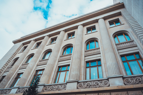 Low Angle Architectural Exterior of Modern Low Rise Building in Classical Style with Large Windows, Decorative Stone Scroll Work and Pillars, Framed Against Blue Sky with Heavy White Clouds - Photo, Image