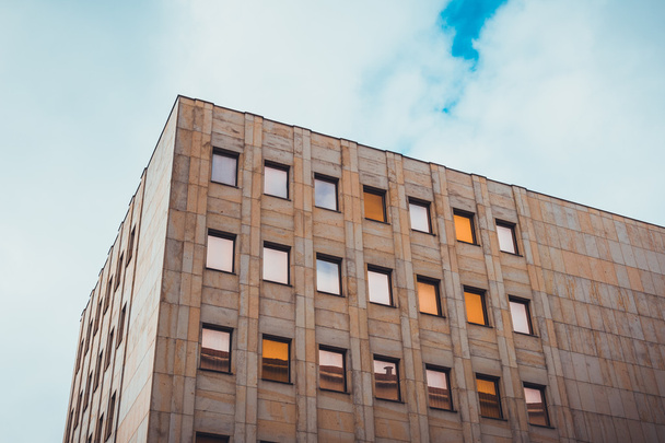 Низкоугловой архитектурный внешний вид современного неописуемого здания с коричневым каменным фасадом и бронзовым цветным отражающим окном, обрамленным голубым небом с тяжелыми белыми облаками
 - Фото, изображение
