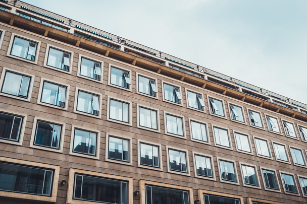 Exterior arquitectónico de bajo ángulo del edificio de oficinas comercial moderno o apartamentos residenciales con fachada marrón, grandes ventanas aireadas y balcones en la azotea enmarcados por cielo nublado gris
 - Foto, imagen