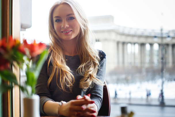 Pretty blonde young woman sitting at the table with vase with Flowers on it in a cafe on the blur background of city - Foto, Bild