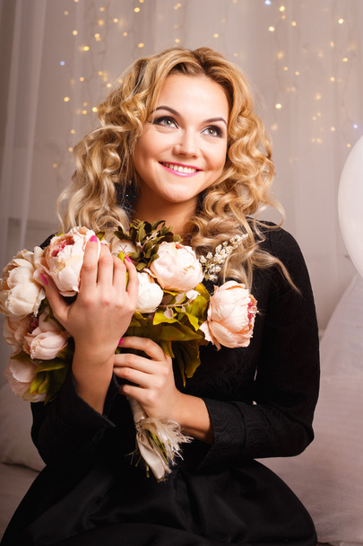 Close-up portrait of beautiful curly young woman sitting on a bed holding bouquet of flowers. Beauty fashion portrait - Fotografie, Obrázek