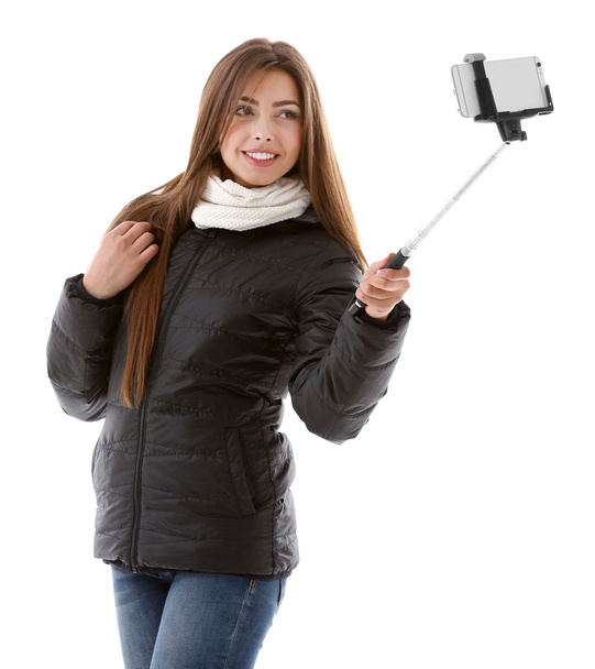 Young attractive girl in winter clothing making photo by her self with mobile phone, isolated on white - Foto, Imagem