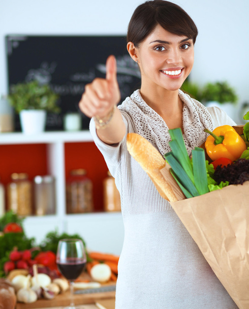Młoda kobieta trzyma torbę na zakupy z warzywami Stojąc w kuchni i pokazując ok - Zdjęcie, obraz