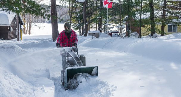 Um homem com roupas de trabalho, chapéu e luvas, operando um lança-neve / ventilador em um dia de inverno no Canadá
. - Foto, Imagem
