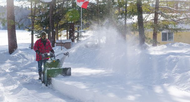 Un hombre en ropa de trabajo, sombrero y guantes, operando un lanzador de nieve / soplador en un día de invierno en Canadá
. - Foto, imagen