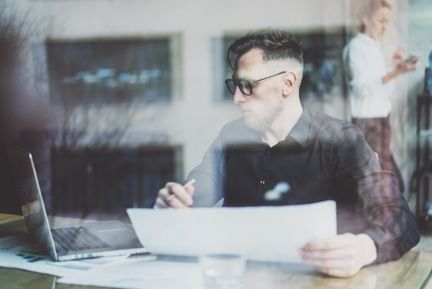 Bearded adult businessman working on modern urban cafe. Photo trough window. Man wearing black shirt and looking laptop, holding papers pencil. Horizontal, film effect, bokeh. - Фото, изображение