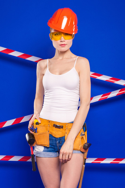 Bouwer meisje op een blauwe achtergrond met een professionele bouwhulpmiddel in een beschermende helm - Foto, afbeelding