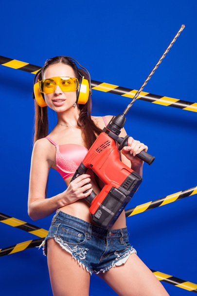 sexy Brünette Bauarbeiter auf blauem Hintergrund mit einem elektrischen Werkzeug in den Händen von - Foto, Bild