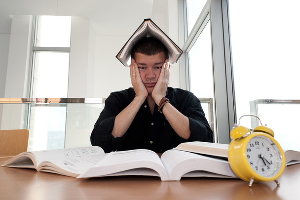 Closeup portrait of young man surrounded by tons of books, alarm clock, stressed from project deadline, study, exams. Negative emotion facial expression feelings, body language - Photo, Image