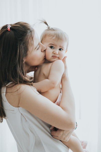Beautiful mother portrait playing with her cute little girl in a white room in the morning - Photo, Image