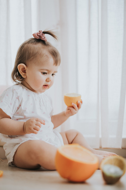 Cute little baby girl on the table in the kitchen eats citrus fruits full of vitamins and makes funny cute faces - Фото, изображение
