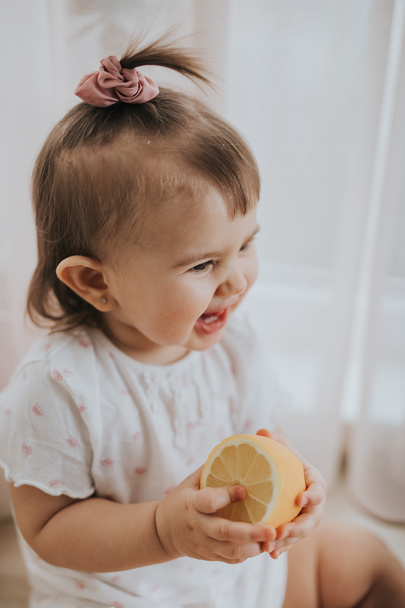 Cute little baby girl on the table in the kitchen eats citrus fruits full of vitamins and makes funny cute faces - Photo, Image